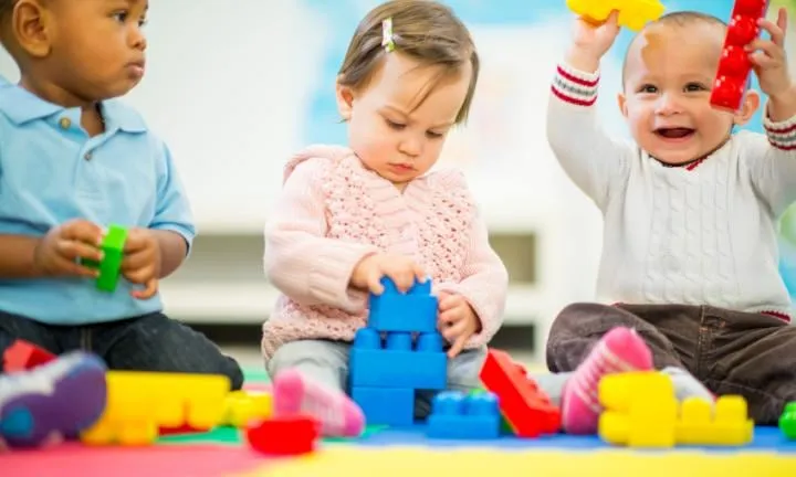 Three toddlers sitting on a play mat stacking and lifting colourful oversized building blocks