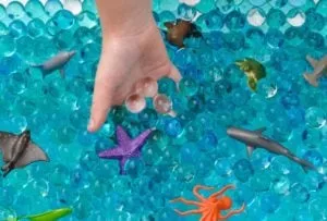 Child's hand in a sensory bin of blue water beads with toy sea animals